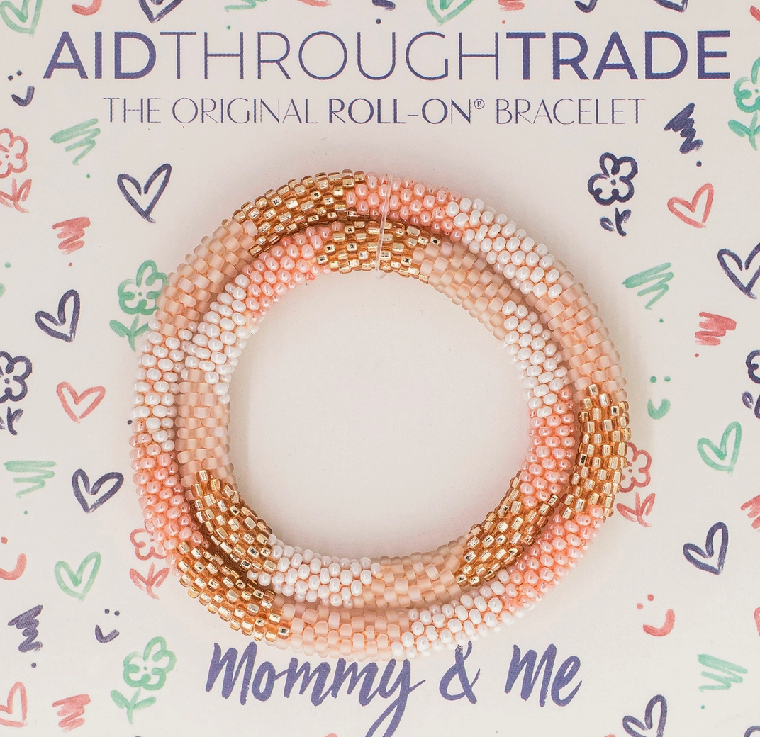 Vor einem lebhaften Hintergrund, der mit kleinen Kritzeleien verziert ist, ist ein Set Perlenarmbänder in Rosa-, Gold- und Weißtönen zu sehen. Der Text lautet „Aid Through Trade – Armbänder Mommy & Me“ und hebt faireren Handel hervor. Die Armbänder sind aus hochwertigen Glasperlen gefertigt.