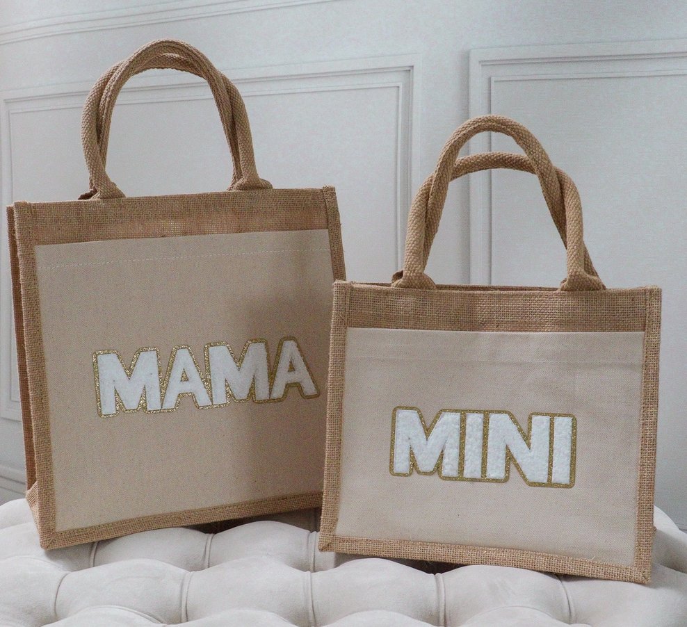 Vor einer weißen Wand sind zwei beigefarbene Tragetaschen mit Seilhenkeln zu sehen. Die größere Tasche trägt das Wort „MAMA“ in goldbesetzten Buchstaben, während die kleinere Tasche das Wort „MINI“ in ähnlichen Buchstaben trägt. Diese nachhaltig gefertigten Kiss Couture – Jutebeutel Mama & Mini von Kiss Couture sind in unserem Kinderartikelgeschäft erhältlich und stehen auf einer getufteten Oberfläche.