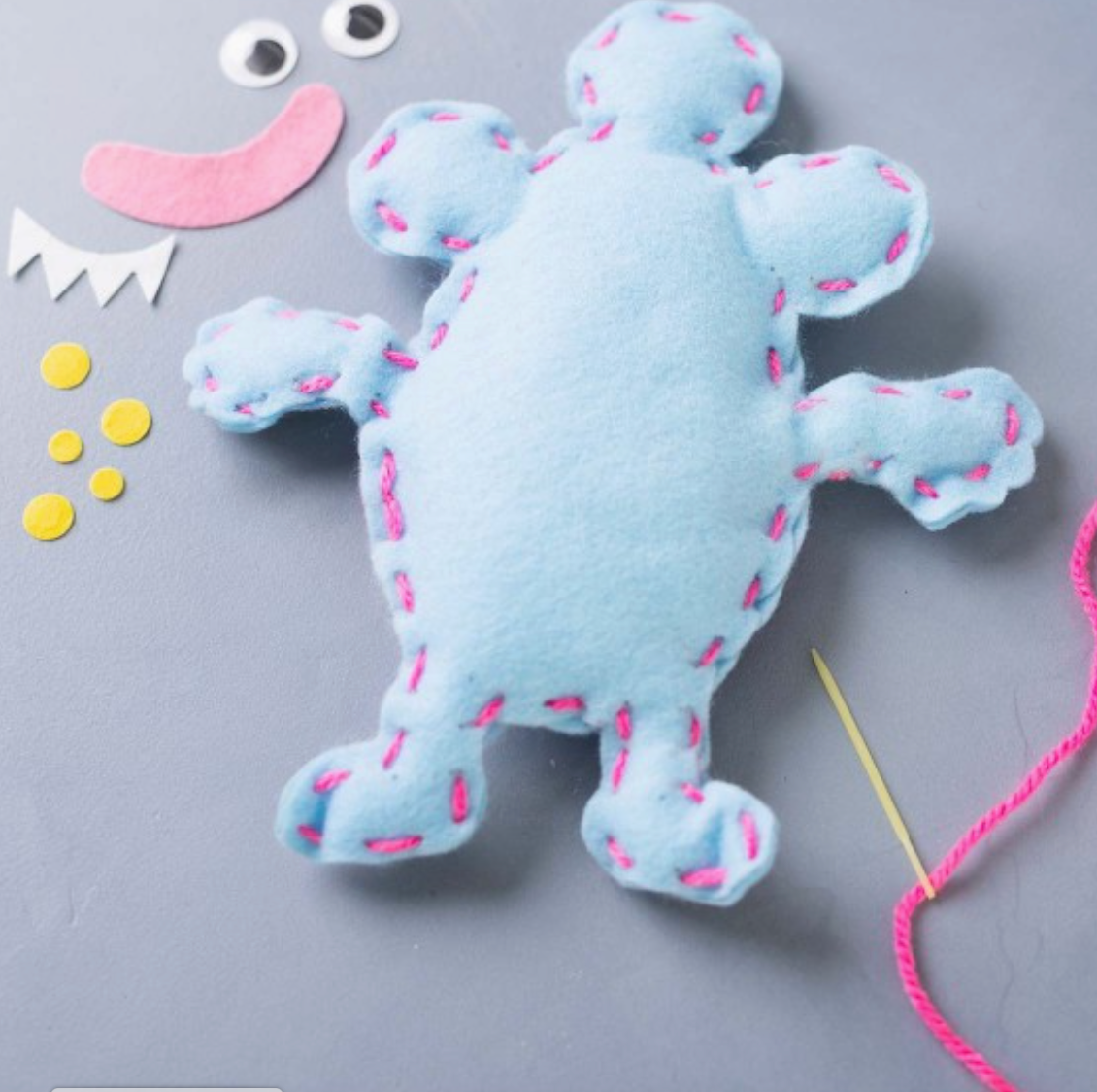 Ein von **Creativ Company - DIY Nähset für Kinder - Monster aus Filz** handgefertigtes blaues Stoffspielzeug mit sechs runden Vorsprüngen ist mit rosa Garn genäht. Darum herum befinden sich ein Paar Filzaugen, ein rosa Filzlächeln, kleine gelbe Filzkreise, eine Nadel und ein Stück rosa Garn auf grauem Untergrund – ideal zum leichten Basteln mit Kindern.