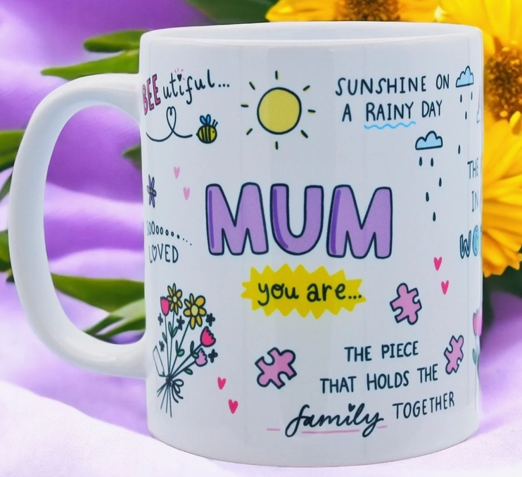 Eine weiße Tasse mit farbenfrohen Illustrationen und herzlichen Botschaften. Auffälliger Text in Pink lautet „MAMA“, umgeben von fröhlichen Zeichnungen von Sonne, Bienen, Blumen und Puzzleteilen. Perfekt als Dekoration fürs Kinderzimmer oder als Ergänzung zu Ihrer Einrichtung fürs Kinderzimmer. Zu den Sätzen gehören „Sonnenschein an einem regnerischen Tag“, „Geliebt“ und „Das Stück, das die Familie zusammenhält“. Dies ist das Feel Good Gift – Tasse Mama von Feel Good Gift.
