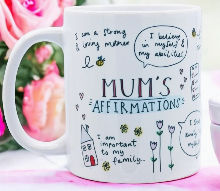 Die Feel Good Gift - Tasse Mama von Feel Good Gift ist mit Aussagen wie „Ich bin eine starke und liebevolle Mutter“ und „Ich glaube an mich und meine Fähigkeiten“ verziert. Die Tasse zeigt farbenfrohe Kritzeleien von Blumen, einem Haus und einer Biene und verleiht jedem Kinderzimmer Charme. Im Hintergrund sind rosa Blumen verschwommen.