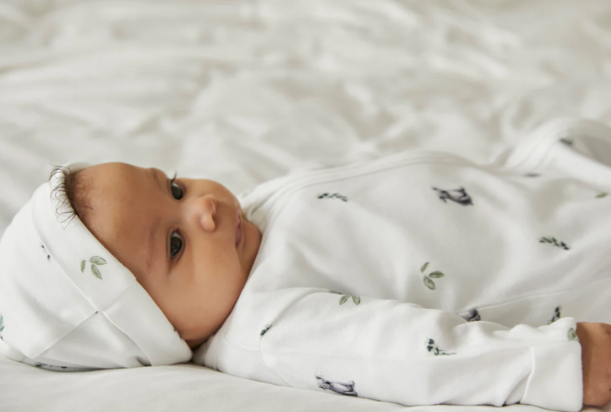 Ein Baby liegt auf einer weißen Decke, trägt einen weißen Strampler und eine passende Rosa & Blue - Mütze von Rosa & Blue, verziert mit kleinen Prints von Blättern und Tieren. Das nachhaltige Outfit ist aus GOTS-zertifizierten Materialien gefertigt. Das Baby schaut zur Seite und zeigt einen ruhigen Gesichtsausdruck.