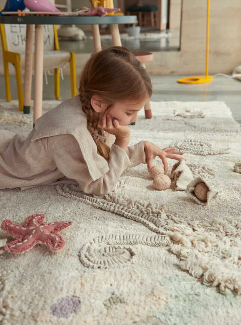 Ein junges Mädchen mit geflochtenen Haaren liegt auf dem Bauch auf einem strukturierten Teppich in einem wunderschön dekorierten Kinderzimmer und spielt konzentriert mit Stofftieren. Sie trägt ein helles Langarmshirt. Im Hintergrund sind verschwommene Möbelstücke und eine Stehlampe zu sehen, die den Lorena Canals - Seabed Spielteppich mit Spielzubehör von Lorena Canals hervorheben.