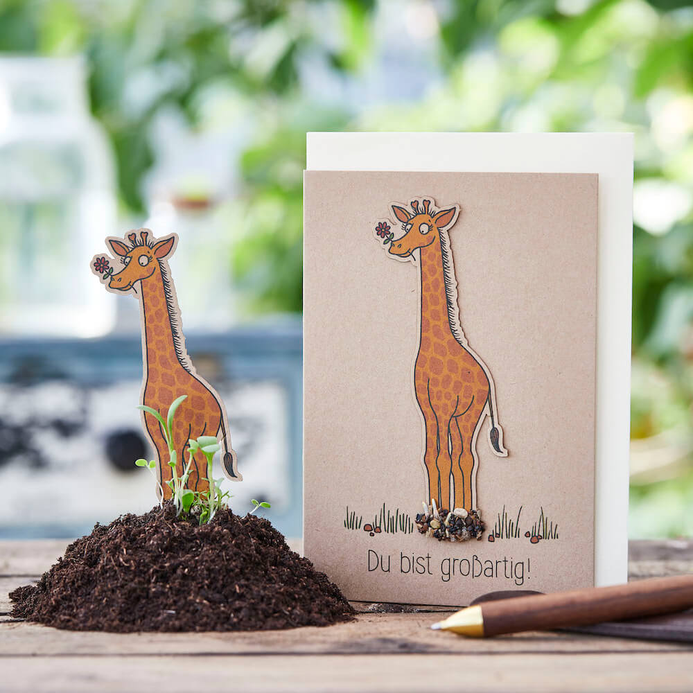 Eine Saatsteckerkarte „Du bist großartig!“ von Die Stadtgärnter mit einer Giraffenillustration steht auf einem Tisch neben einem kleinen Erdhaufen, in den eine Giraffe aus Papier eingelegt ist. Auf der Karte steht „Du bist großartig!“, was übersetzt „Du bist großartig!“ bedeutet. Neben der Karte liegt ein Stift, perfekt zum Basteln einer Geburtstagskarte inmitten des verschwommenen Grüns.