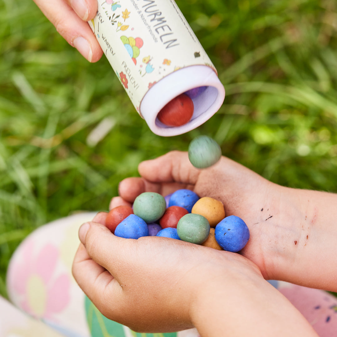 Nahaufnahme der Hände eines Kindes, das bunte Blumenmurmeln in Dosen von Die Stadtgärnter hält, die aus einer Tube von Die Stadtgärnter gegossen werden. Die Hände und Samenkugeln sind im Fokus, mit einem grünen Grashintergrund. Die Samenkugeln haben verschiedene Farben, darunter blau, grün und rot. Das Kind trägt bunt gemusterte Hosen.