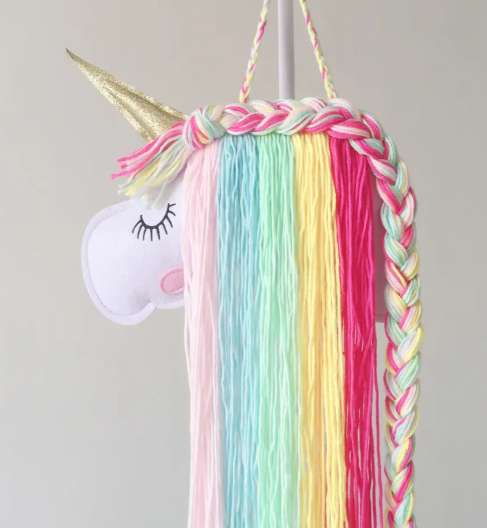 Ein Artilady - Einhorn-Haarspangenhalter aus Filz und Garn, perfekt für ein Babyzimmer-Tierthema. Das majestätische Einhorn hat ein goldenes Horn, geschlossene Augen mit schwarzen Wimpern und eine pastellfarbene geflochtene Mähne in den Farben Rosa, Blau, Gelb und Grün vor einem schlichten hellgrauen Hintergrund.