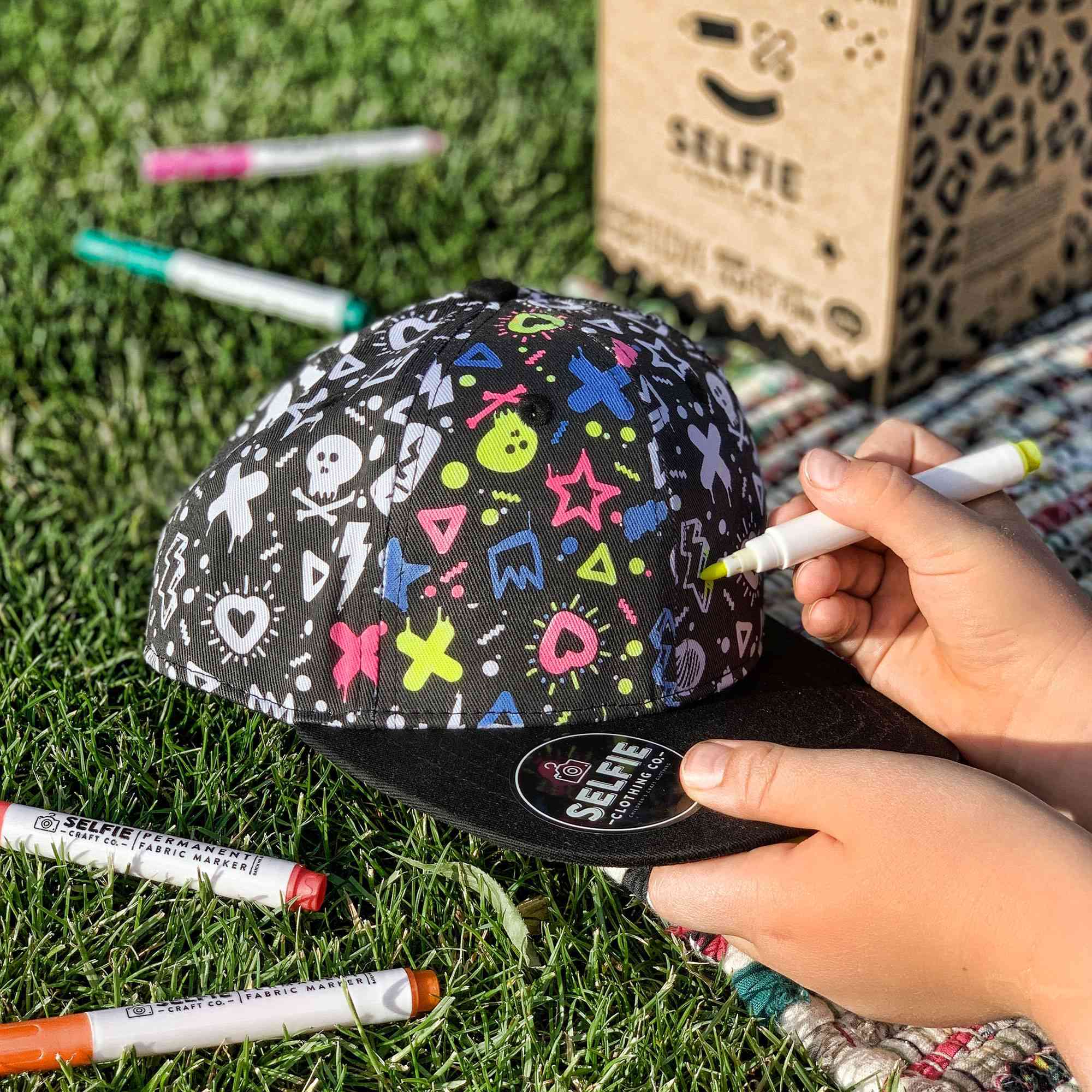 Nahaufnahme einer Person, die eine Selfie Craft Co. - Graffiti Colour im Snapback Kappe mit bunten Markern von Selfie Craft verziert. Die Kappe zeigt verspielte Designs wie Herzen, Sterne und verschiedene Symbole. Im Hintergrund sind zusätzliche Marker und eine Box mit der Aufschrift „Selfie“ zu sehen, alles auf Gras platziert.