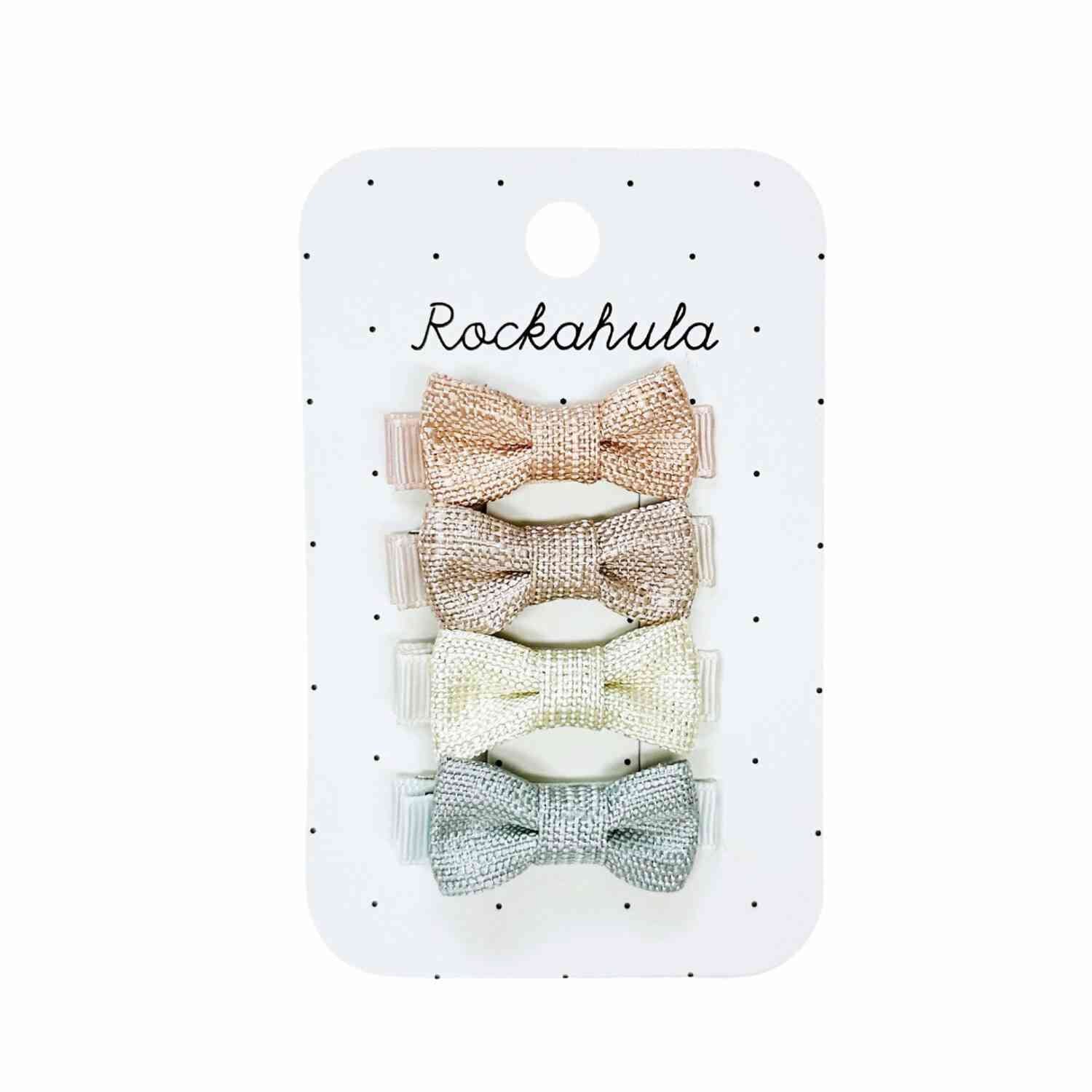 Auf einer Karte mit der Aufschrift „Rockahula“ befinden sich vier glitzernde Rockahula – Schimmernde Mini-Schleifenclips, perfekt für kleine Prinzessinnen. Von oben nach unten: Rosa, Gold, Silber und Mintgrün. An jeder Schleife ist eine Haarspange befestigt und der Kartenhintergrund ist weiß mit kleinen schwarzen Punkten.