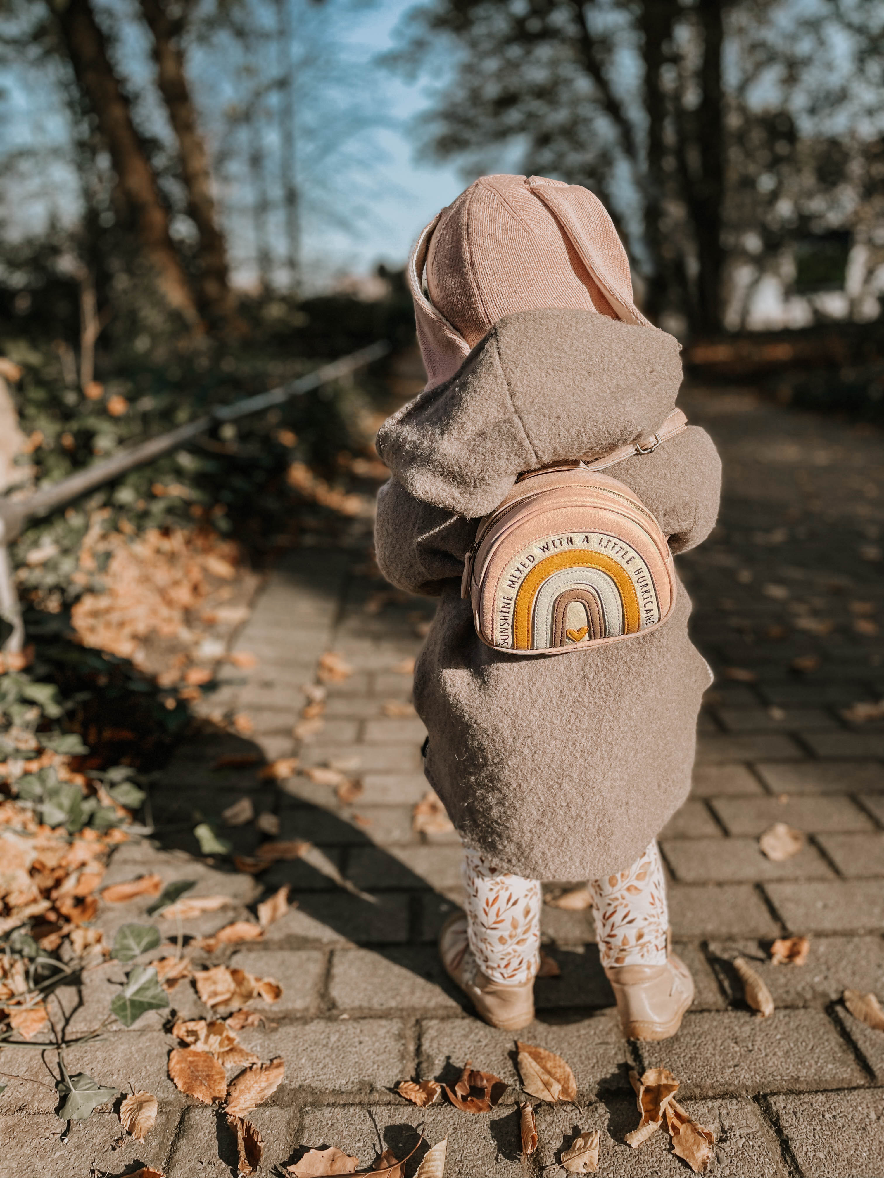 Ein kleines Kind mit einer rosa Kapuze und einem grauen Mantel mit Bärenohren steht auf einem mit Blättern übersäten Weg. Das Kind trägt einen Little Who - Rucksack Regenbogen Gerti mit den Worten „dreaming with a little...“ darauf, verziert mit reflektierenden Aufklebern vom örtlichen Kinderartikelgeschäft. Im Hintergrund sind Bäume und Grün zu sehen.