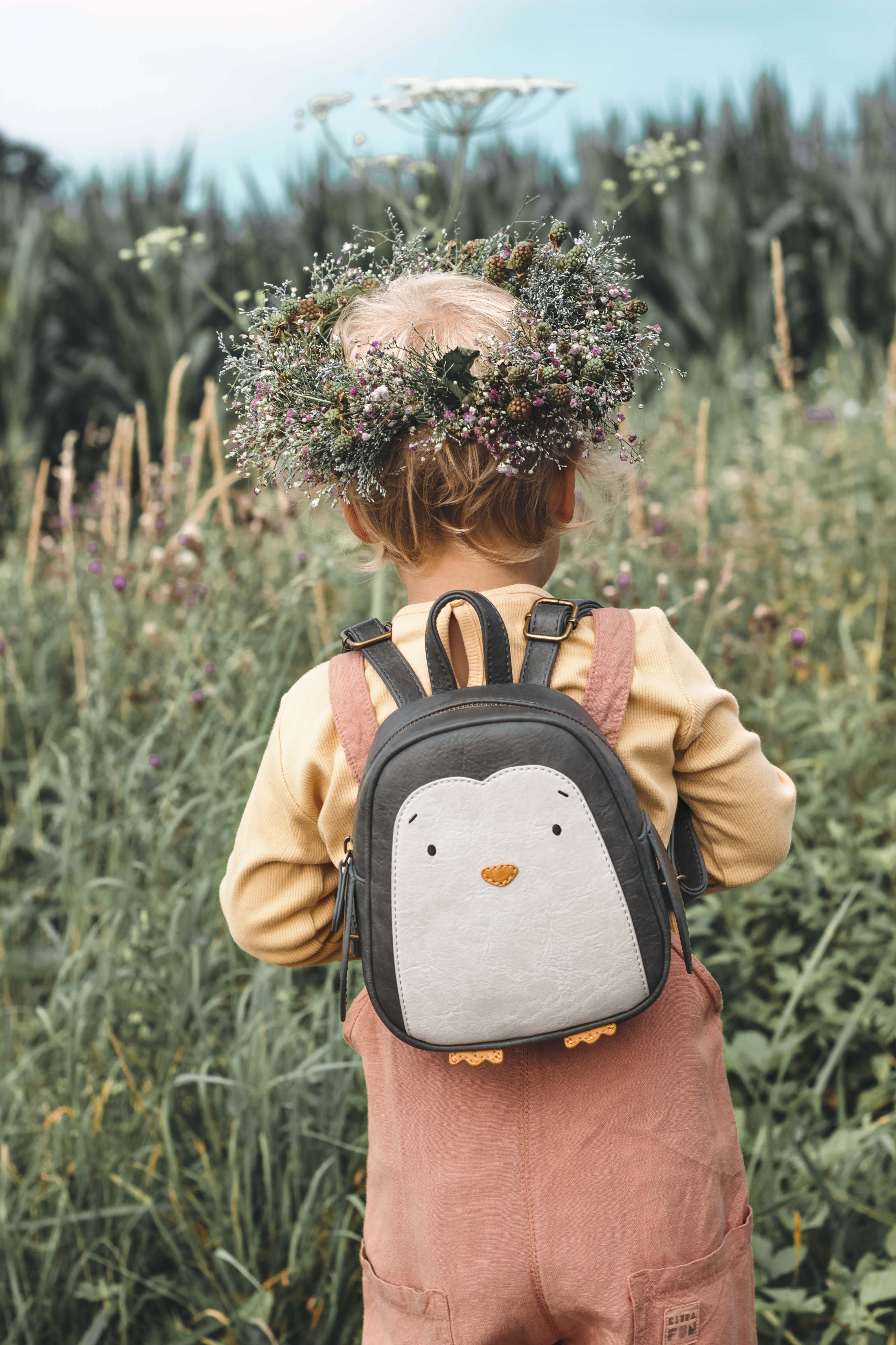 Ein kleines Kind mit kurzen blonden Haaren und einem Blumenkranz steht mit abgewandtem Blick auf einer Wiese. Das Kind trägt ein gelbes Hemd, einen rosa Overall und einen Little Who - Rucksack Pinguin Lilo von Little Who, der mit reflektierenden Aufklebern verziert ist. Die Wiese ist üppig mit grünem Gras und Wildblumen bewachsen.