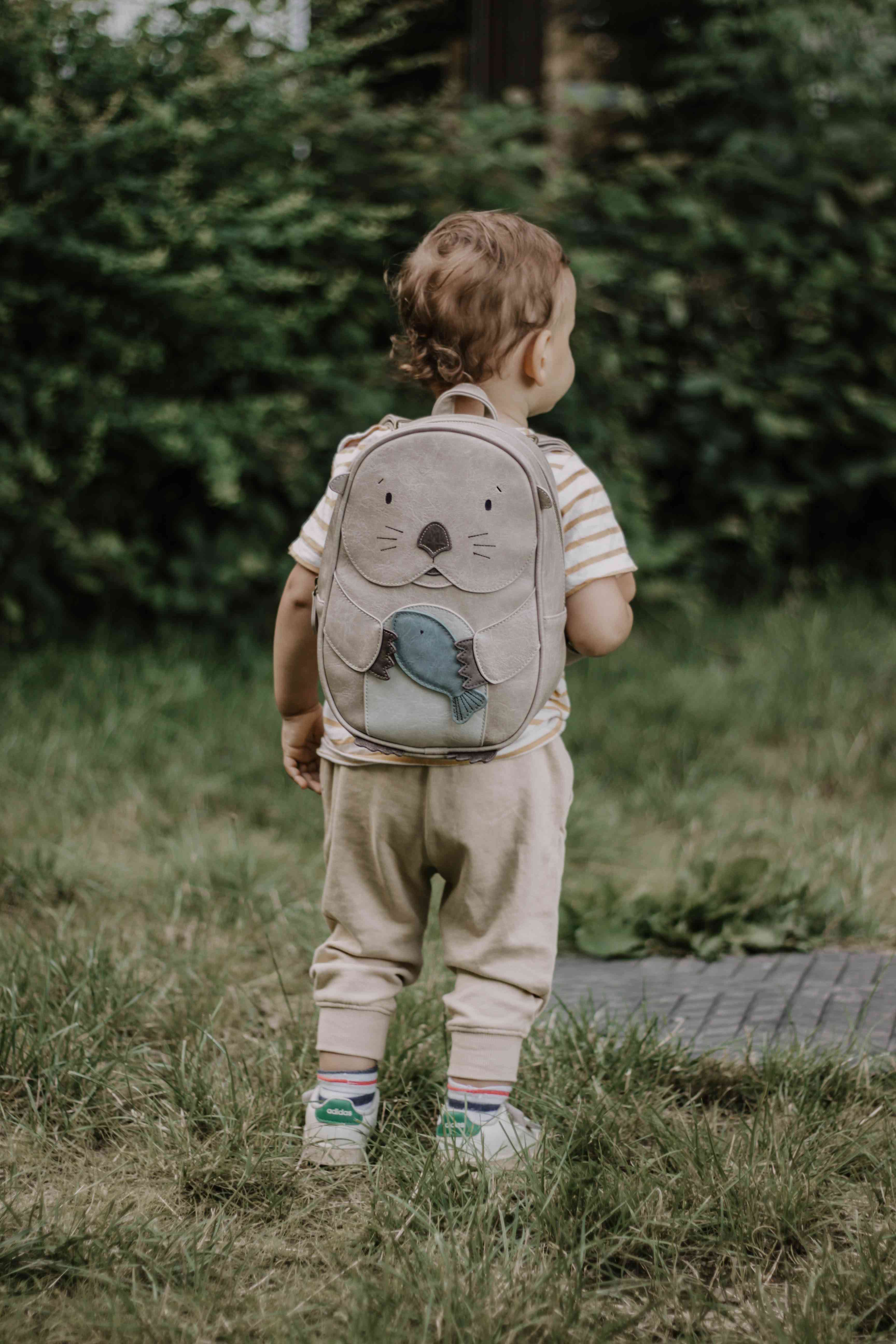 Ein kleines Kind mit lockigem Haar steht auf dem Gras. Es trägt ein creme- und beige gestreiftes Hemd, beige Hosen und bunte Socken mit weißen Schuhen. Das Kind trägt den Little Who - Rucksack Otter Mathilda von Little Who – einen grauen Rucksack mit einem süßen Otter-Design mit einem blauen Fisch – erhältlich bei Ihrem örtlichen Kinderartikelgeschäft. Im Hintergrund ist üppiges grünes Laub zu sehen.