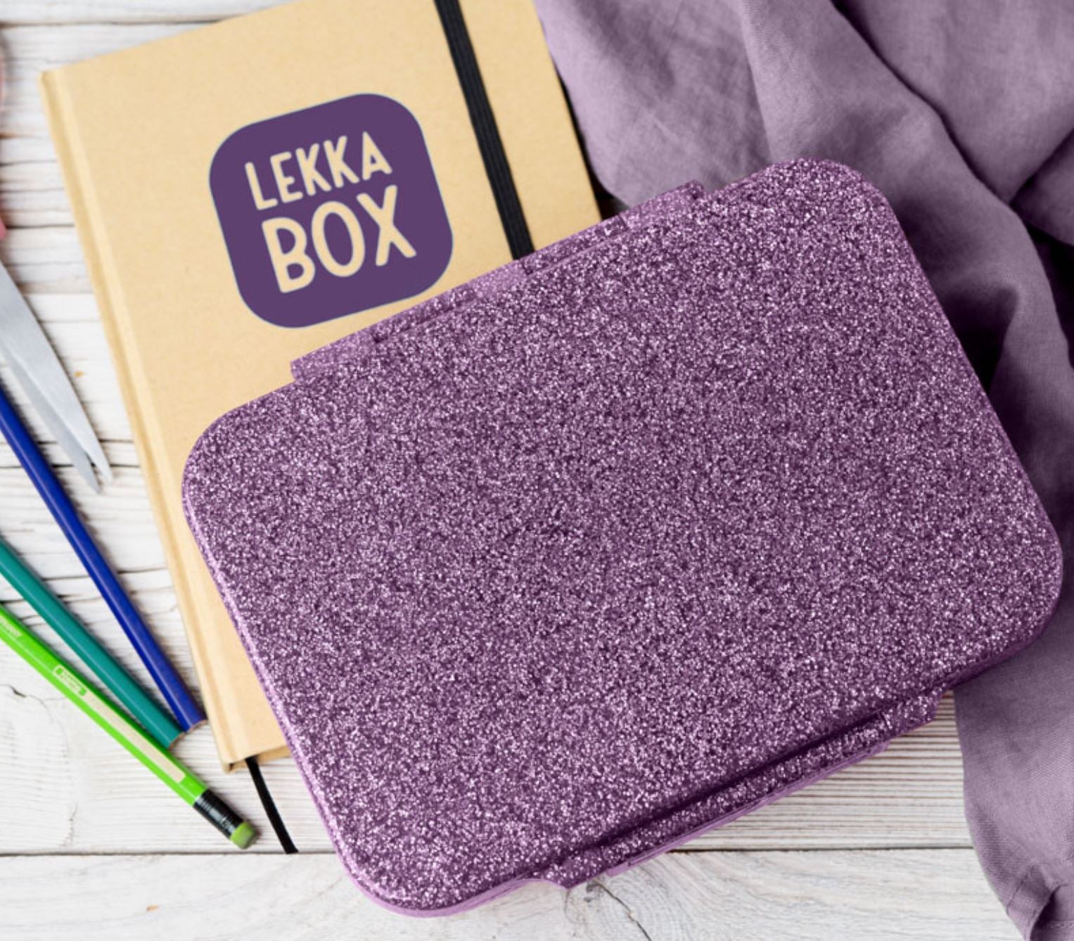 Eine glitzernde lila Lunchbox steht auf einem Tisch neben einem beigen Notizbuch mit der Aufschrift „LEKKA BOX“ auf dem Umschlag. Daneben liegen mehrere Buntstifte und ein lila Tuch, das die Lekkabox – Lunchbox Glamour von Lekkabox – ergänzt. Der Hintergrund besteht aus einer weißen Holzoberfläche.