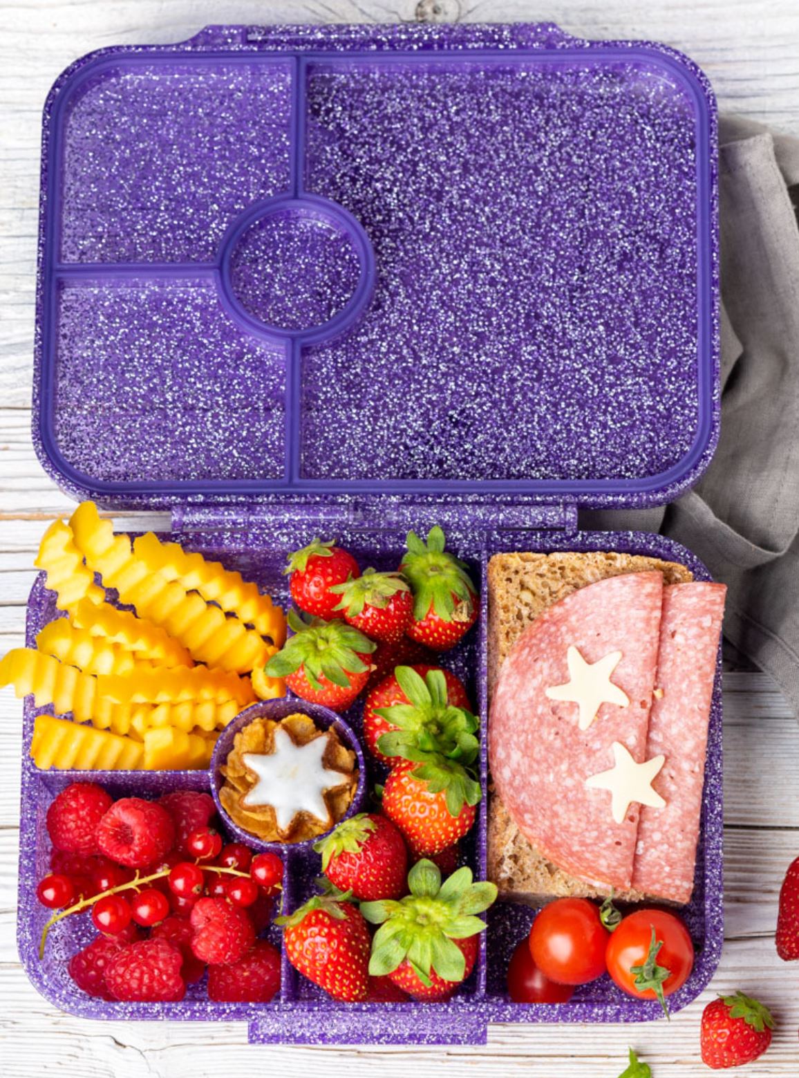 Eine violette Lunchbox mit mehreren Fächern, die Himbeeren, Curly Fries, rote Johannisbeeren, Erdbeeren, Kirschtomaten, ein kleines sternförmiges Dessert und ein Salami-Käse-Sandwich mit zwei sternförmigen Käsescheiben enthält. Diese Lekkabox - Lunchbox Glamour von Lekkabox ist perfekt zum Verpacken einer köstlichen Mahlzeit.