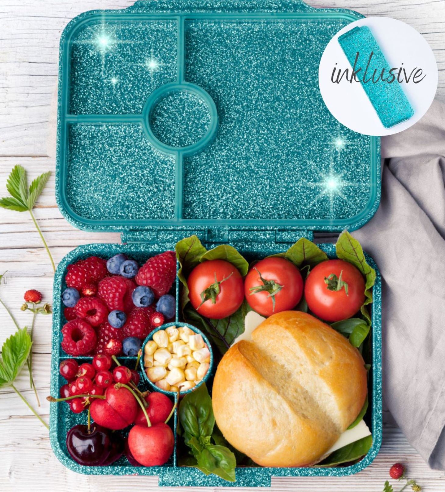 Eine türkise Lekkabox - Lunchbox Glamour von Lekkabox mit mehreren Fächern, die Folgendes enthalten: Kirschtomaten, Spinatblätter, ein Sandwich mit rundem Brötchen, gemischte Beeren (Erdbeeren, Himbeeren, Heidelbeeren), Kirschen und eine kleine Tasse Maiskörner. Ein helltürkisfarbener Stoff liegt daneben. Perfekt als Znünibox für den Kindergarten.