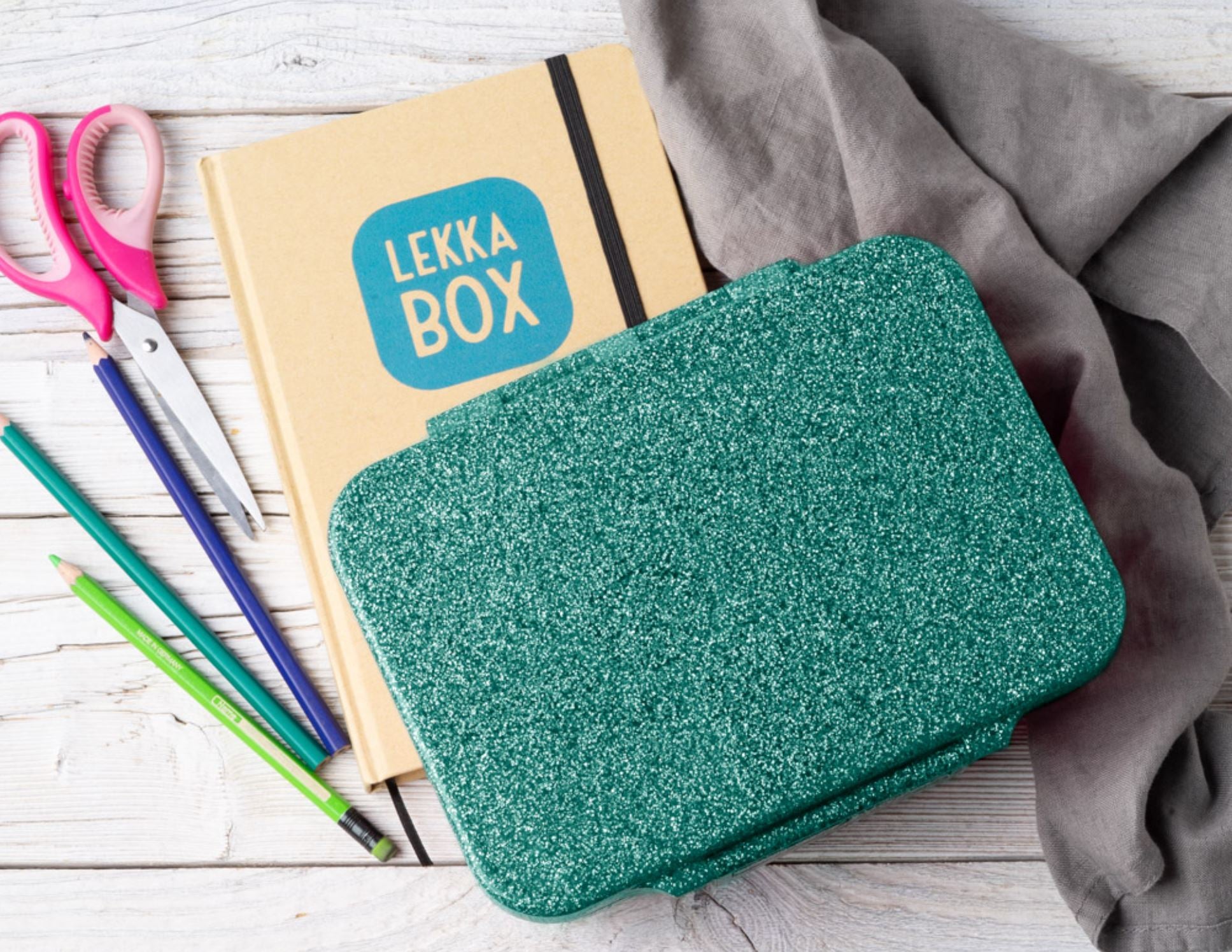 Eine glitzernde grüne Lekkabox – Lunchbox Glamour liegt auf einer Holzoberfläche neben einem braunen Notizbuch mit der Aufschrift „Lekkabox“ auf dem Umschlag. Daneben liegen eine rosa Schere, drei Buntstifte (blau, grün und schwarz) und ein zerknittertes graues Tuch. Diese Lekkabox – Lunchbox Glamour verleiht der Mittagspause jedes Kindes Charme.