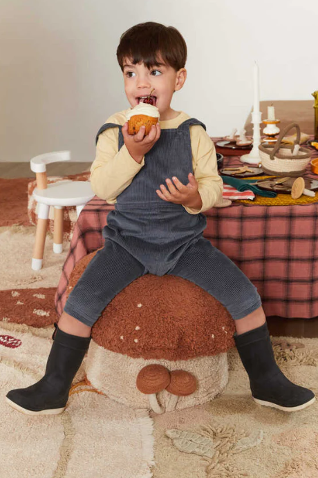 Ein kleines Kind sitzt auf einem Sitzkissen Boletus von Lorena Canals und genießt einen Muffin. Mit einem dunklen Overall, einem hellen Langarmshirt und schwarzen Stiefeln fühlt sich die Szene wie aus einem bezaubernden Kinderzimmer an. Hinter ihnen steht ein Tisch mit einer karierten Tischdecke, auf dem Snacks und eine Kerze liegen, während links ein kleiner Stuhl steht.