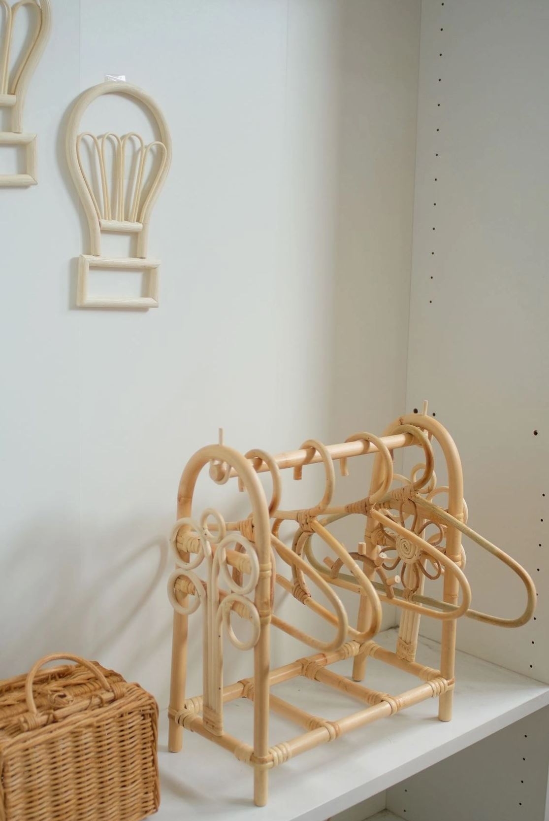 Ein beiges Zimmer, dekoriert mit Holzelementen. Links steht ein Rattankorb auf einem Regal. Darüber hängen zwei Rattan-Wandbehänge in Form von Luftballons. In der Mitte hält der CindyxHanna - Puppen Kleiderständer von CindyxHanna mehrere Holzbügel mit kreisförmigen Akzenten – perfekt für eine nachhaltige Kinderzimmerdekoration.