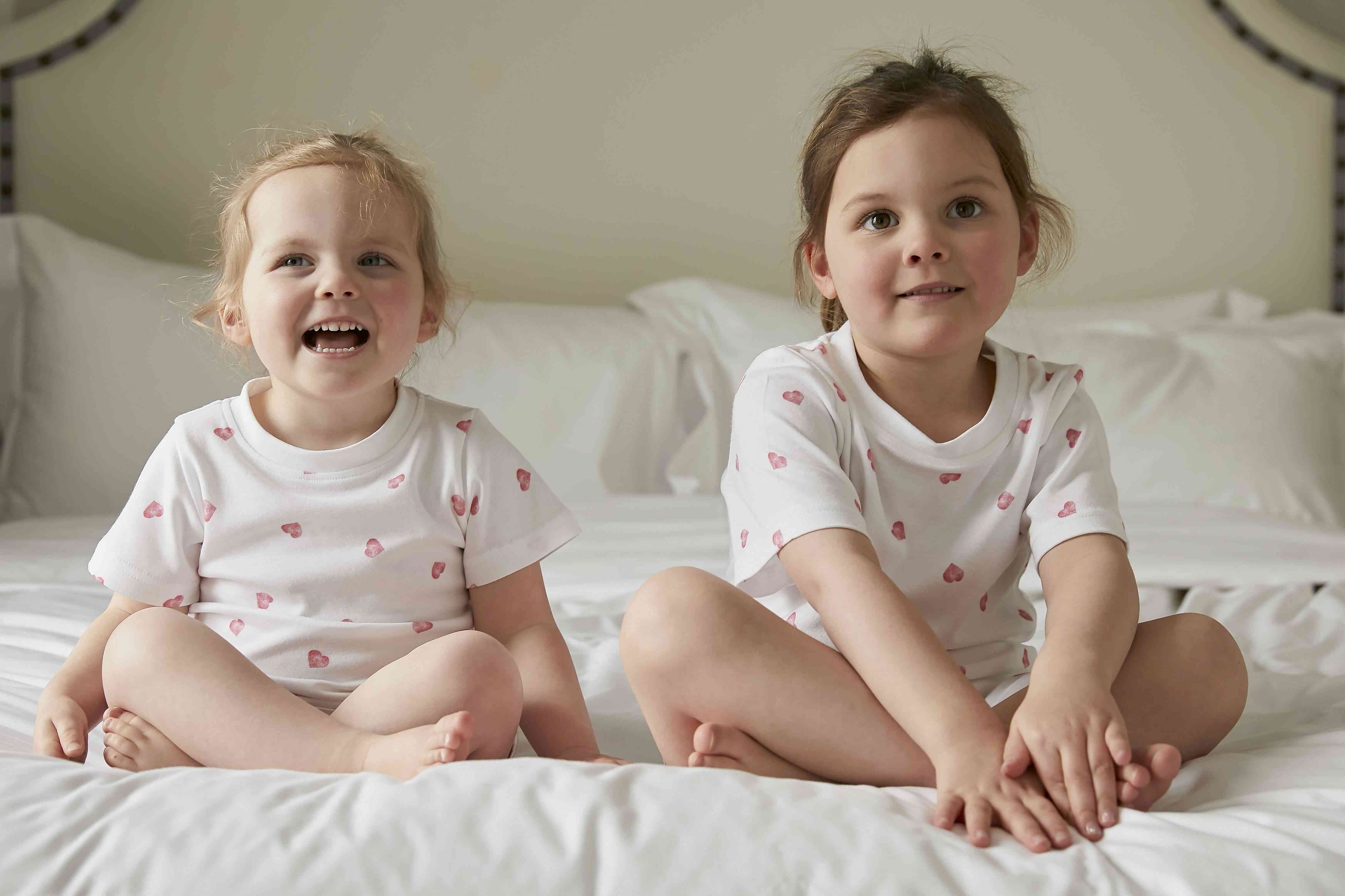 Zwei kleine Kinder sitzen auf einem Bett, beide tragen Rosa & Blue - Pyjamas kurzarm von Rosa & Blue aus Bambus und Bio-Baumwolle, verziert mit rosa Herzmustern. Das Kind links lächelt breit, während das Kind rechts leicht zur Seite blickt. Sie sitzen im Schneidersitz auf einer weißen Bettdecke mit Kissen dahinter.