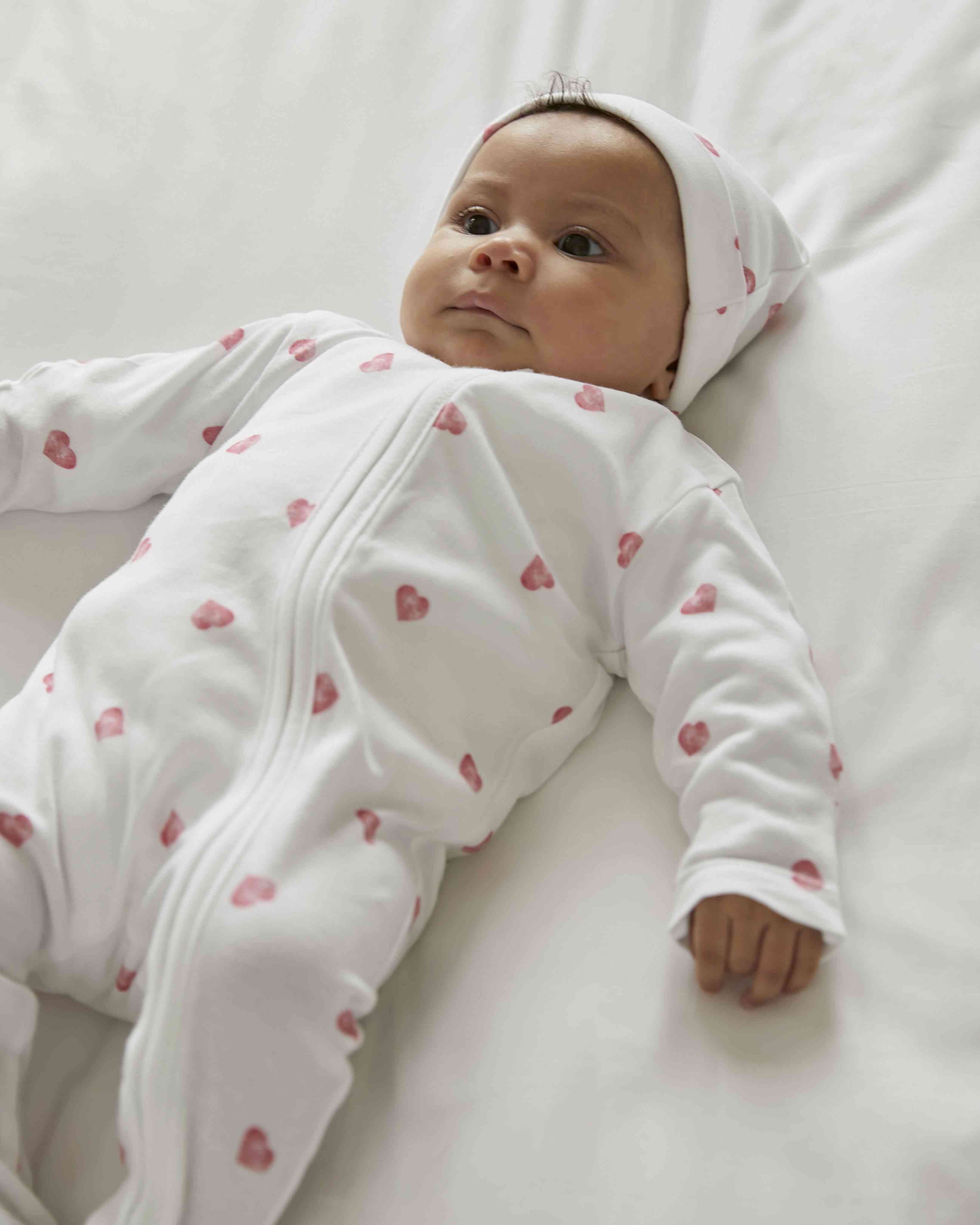 Ein Baby in einem weißen Strampler und einer passenden Mütze mit rosa Herzmuster aus der Rosa & Blue - Schlafstrampler langarm-Kollektion von Rosa und Blue liegt auf einem weißen Laken. Das Baby hat einen ruhigen Gesichtsausdruck und blickt nach oben.