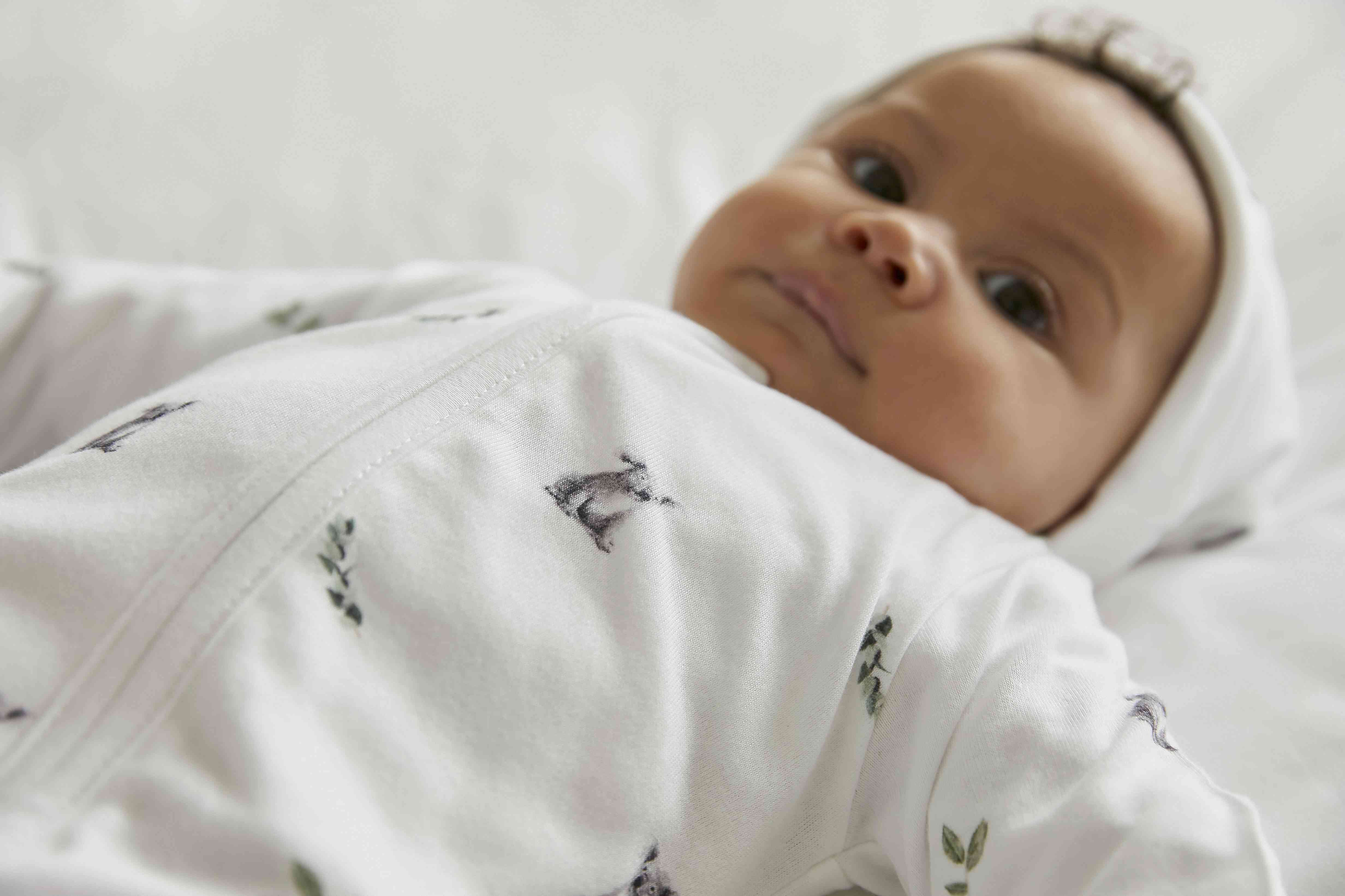 Ein Baby in einem Rosa & Blue Rosa & Blue - Schlafstrampler Langarm-Strampler mit kleinen Tiermotiven liegt auf einer weichen, weißen Oberfläche. Das Baby blickt mit sanftem Gesichtsausdruck nach oben und trägt eine passende weiße Mütze aus unserer OEKO-TEX zertifizierten Kollektion.