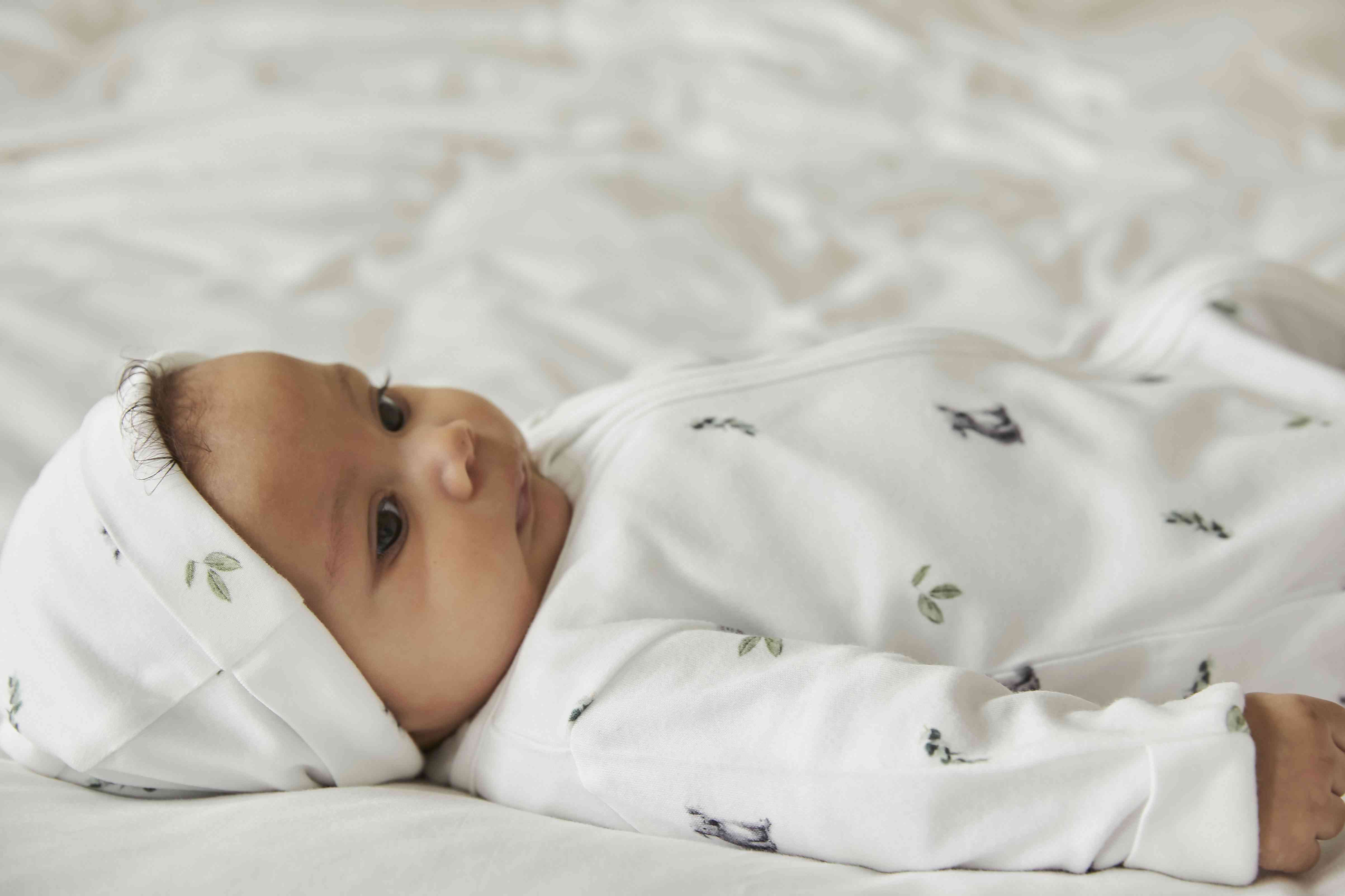 Ein Baby liegt auf einer weißen, plüschigen Oberfläche, trägt einen Rosa & Blue - Schlafstrampler langarm von Rosa & Blue und eine passende Mütze mit Blättermuster und blickt nach links. Das Baby hat einen ruhigen Gesichtsausdruck und ist in kuschelige Bettwäsche gebettet. Der Hintergrund ist sanft mit Weißtönen verwischt.