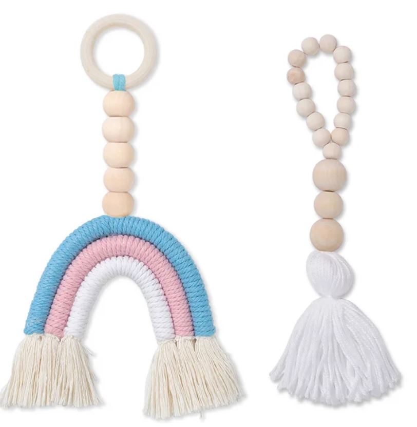 Zwei Makramee-Babyspielzeuge. Das linke Spielzeug hat einen Ring und Holzperlen, die zu einem pastellfarbenen Regenbogen in Creme, Rosa und Blau führen und in Quasten enden. Das rechte Spielzeug hat Holzperlen, die in einer dicken, weißen Quaste enden. Perfekt für Ihr Kinderzimmer und als nachhaltige Kinderprodukte gefertigt, haben beide ein natürliches, handgemachtes Aussehen. Produktname: Artilady - Regenbogen Anhänger Makramee Markenname: Artilady