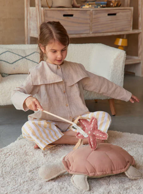 Ein kleines Kind sitzt auf einem Teppich und hält spielerisch einen Zauberstab mit einem sternförmigen rosa Ornament über einem Plüschkissen „Kissen Turtle“ von Lorena Canals. Das Kind trägt ein beiges Oberteil und gestreifte Hosen. Im Hintergrund ist das gemütliche Kinderzimmer mit weißen Holzmöbeln zu sehen.