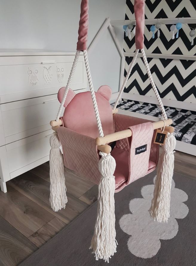 Ein handgefertigter rosa BabyUniquecorn - Kleinkind Swing Bear mit weißer, synthetischer Hanfseile und Quasten hängt in einem modernen Kinderzimmer. Der Sitz hat Bärenohren an der Rückenlehne. Im Hintergrund sind eine weiße Kommode mit Tierzeichnungen, eine schwarz-weiße Wand mit Chevron-Muster und ein grau-weißer Teppich zu sehen.