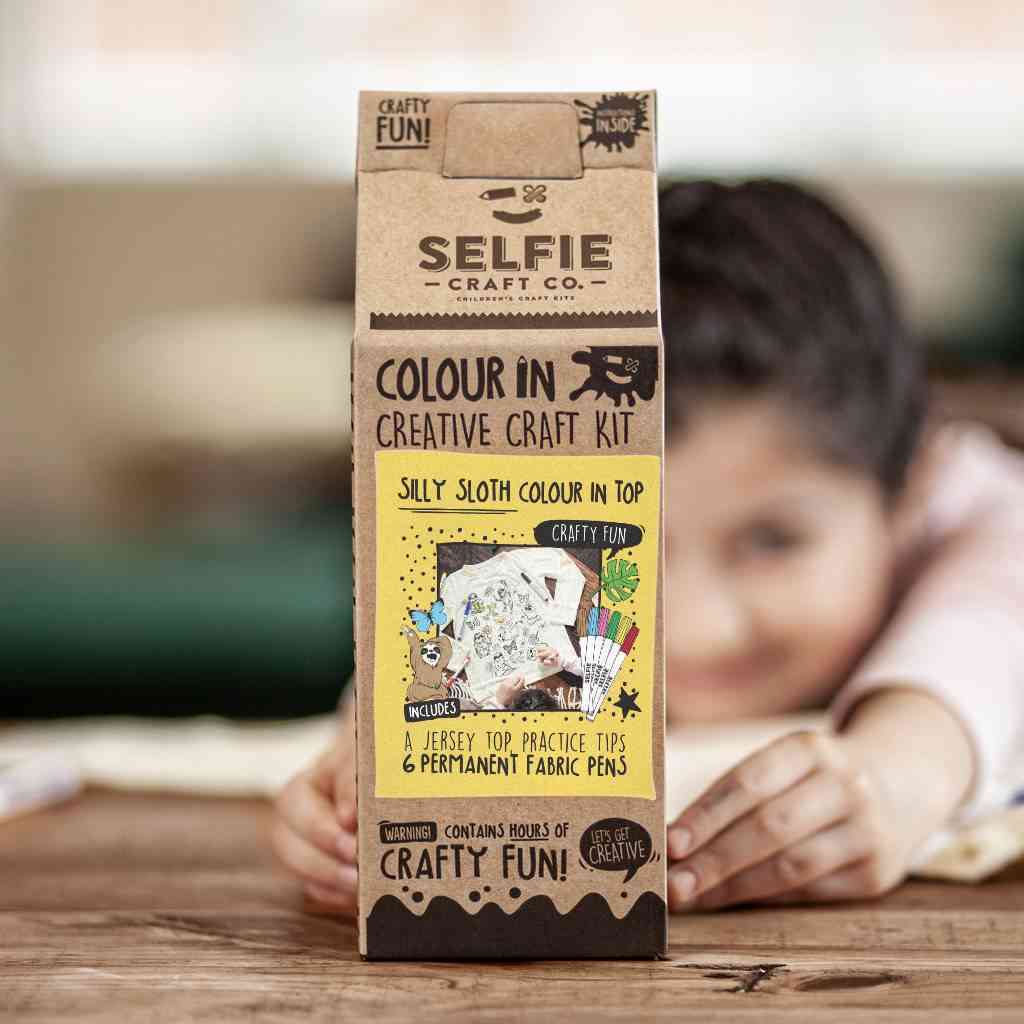 Ein Kind hält eine „Selfie Craft“-Box in der Hand, die ein „Selfie Craft Co. – Faultiere zum Ausmalen“ enthält. Dieses DIY-Set enthält ein Jersey-Oberteil, Übungsspitzen und permanente Stoffstifte. Die Verpackung zeigt verspielte, bastelbezogene Illustrationen und trägt die Aufschrift „Silly Sloth Colour In Top“, perfekt zum leichten Basteln mit Kindern.