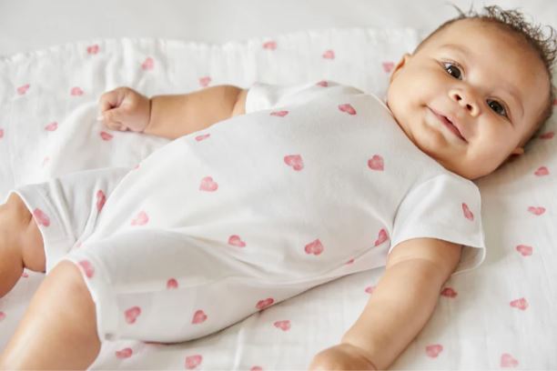 Ein lächelndes Baby liegt auf einer Decke, gekleidet in einen weißen **Rosa & Blue - Strampler kurzarm** mit rosa Herzmuster aus antiallergener Bambus-Bio-Baumwolle. Das Baby hat lockiges Haar und blickt herzlich in die Kamera, was eine herzerwärmende und freudige Szene schafft.