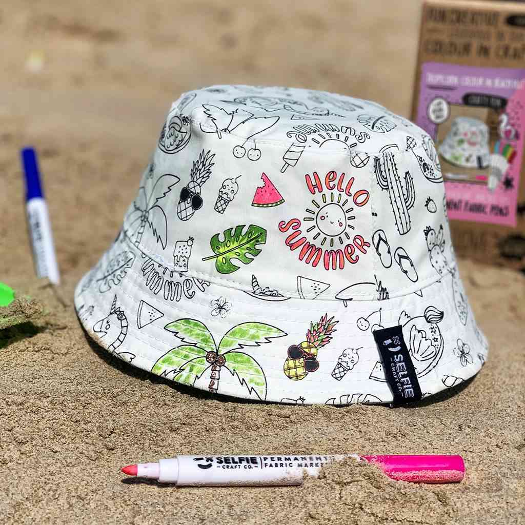 Ein weißer Anglerhut mit verschiedenen sommerlichen Illustrationen und der Aufschrift „Hello Summer“ steht auf sandigem Boden. Daneben liegen farbige Marker und eine Kartonverpackung für den Selfie Craft Co. - Tropicorn Strandhut, die darauf hinweist, dass es sich um ein DIY-Set für kreativen Spaß von Selfie Craft handelt.