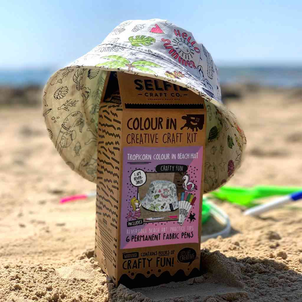 Eine Schachtel mit der Aufschrift „Selfie Craft Co. – Tropicorn Strandhut“ oder „Bastelset 4 Jahre“ steht an einem Sandstrand. Auf der Schachtel liegt ein bunter Sonnenhut. Die Strandszene zeigt im Hintergrund einige Sandspielzeuge, in der Ferne ist eine Meeresküste zu sehen.