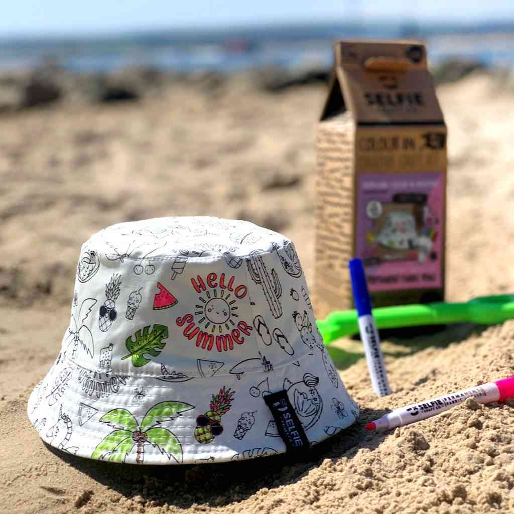 Ein weißer Anglerhut mit bunten Zeichnungen und dem Text „Hello Summer“ liegt auf sandigem Boden. Daneben ein Karton mit der Aufschrift „Selfie Craft Co. – Tropicorn Strandhut“, ein rosa-blauer Marker aus einem Selfie Craft DIY-Set und ein grünes Plastikspielzeug. Im Hintergrund ist ein unscharfer Strand- und Meerblick zu sehen.