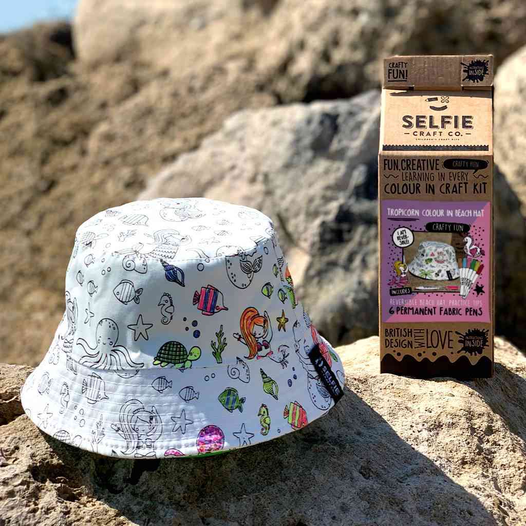 Ein weißer Anglerhut mit bunten, handgezeichneten Meerestieren ist auf einer Felsformation im Freien ausgestellt. In der Nähe steht eine Schachtel mit der Aufschrift „Selfie Craft Co.“, die den Selfie Craft – Tropicorn Strandhut als Teil eines DIY-Sets für leichtes basteln mit kindern bewirbt, das Stoffstifte zum Ausmalen des Hutes enthält. Im Hintergrund sind weitere Felsen und eine verschwommene Landschaft zu sehen.