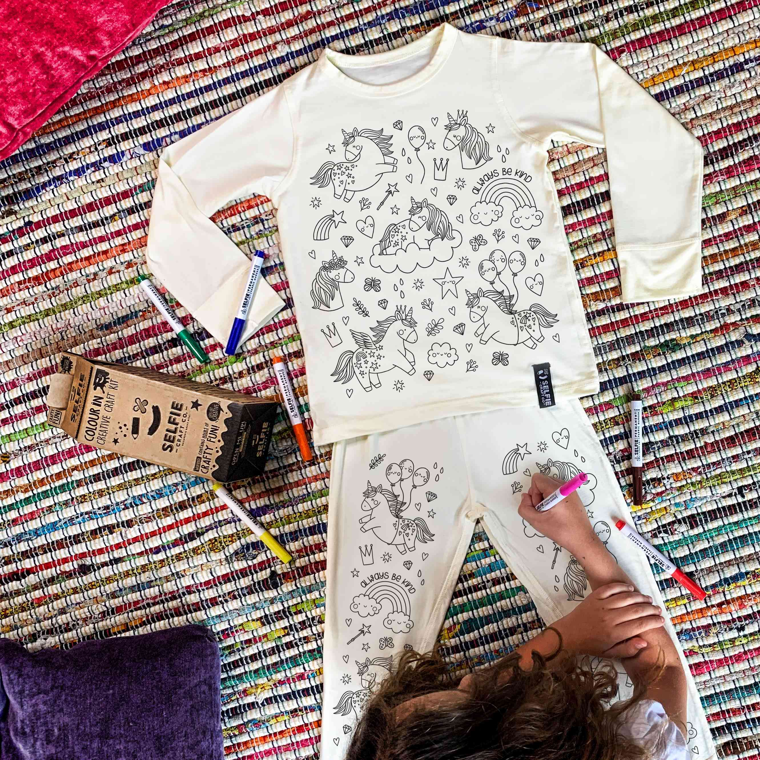 Ein Kind liegt auf einem bunt gestreiften Teppich und malt mit einem Selfie Craft Co. - Pyjama in Einhornfarbe Einhornmotive auf seinen weißen Pyjama. In der Nähe liegen verstreut Buntstifte und eine braune Schachtel mit der Aufschrift „Selfie“. Der Pyjama zeigt verschiedene skurrile Einhornillustrationen, perfekt für kreative Kinder ab 4 Jahren.