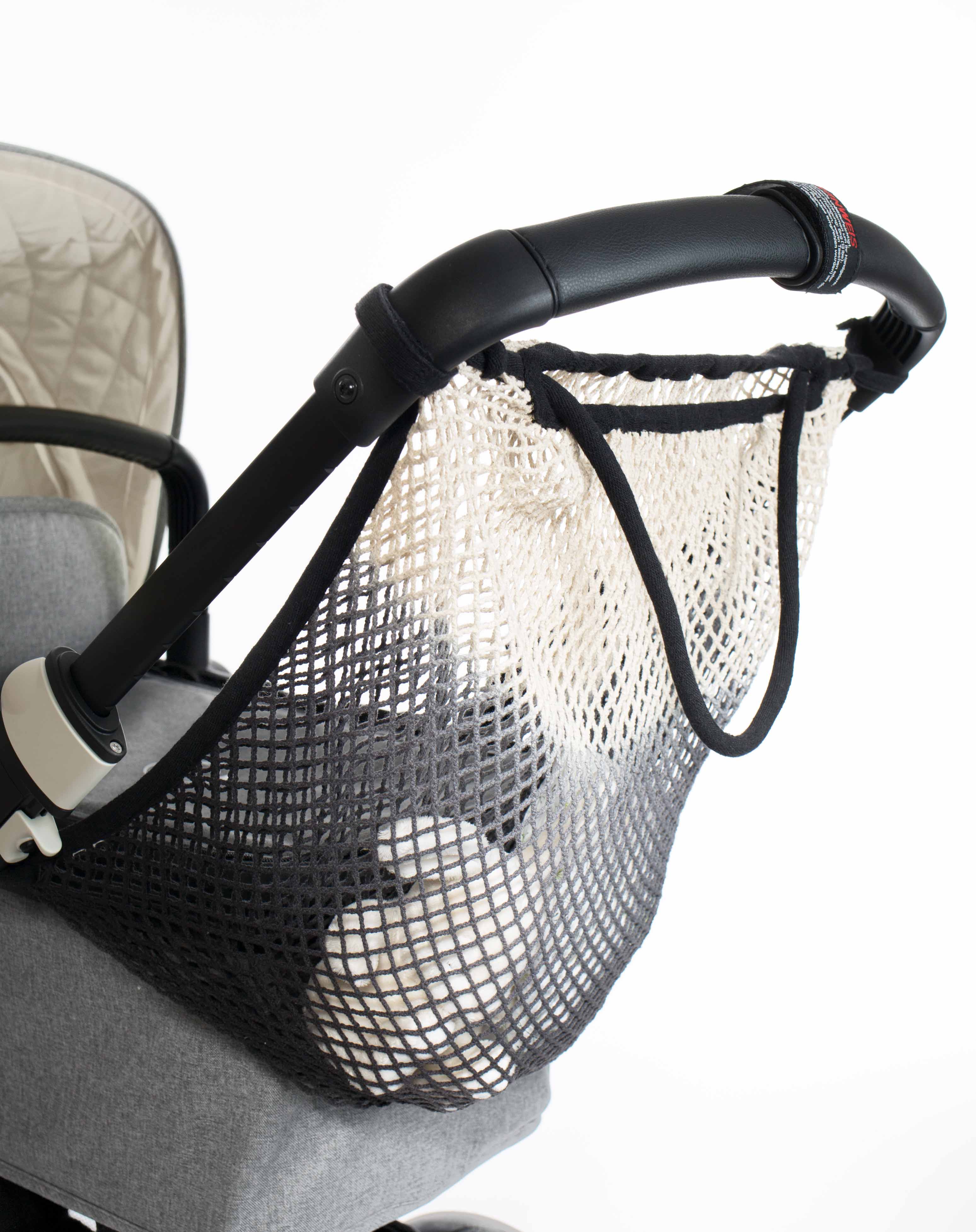 Eine weiß-schwarze Aufbewahrungstasche aus Netzgewebe, das Mara Mea - Kinderwagen Netz - schwarzer Bär von Mara Mea, ist am Griff eines grauen Kinderwagens befestigt und eignet sich perfekt zum Transportieren wichtiger Dinge aus Ihrem Lieblings-Kinderartikelgeschäft. In der Tasche befinden sich ein paar weiße Stofftiere. Der Stoff des Kinderwagens ist gesteppt und der Griff ist schwarz mit einem kreisförmigen rot-weißen Nahtdetail.