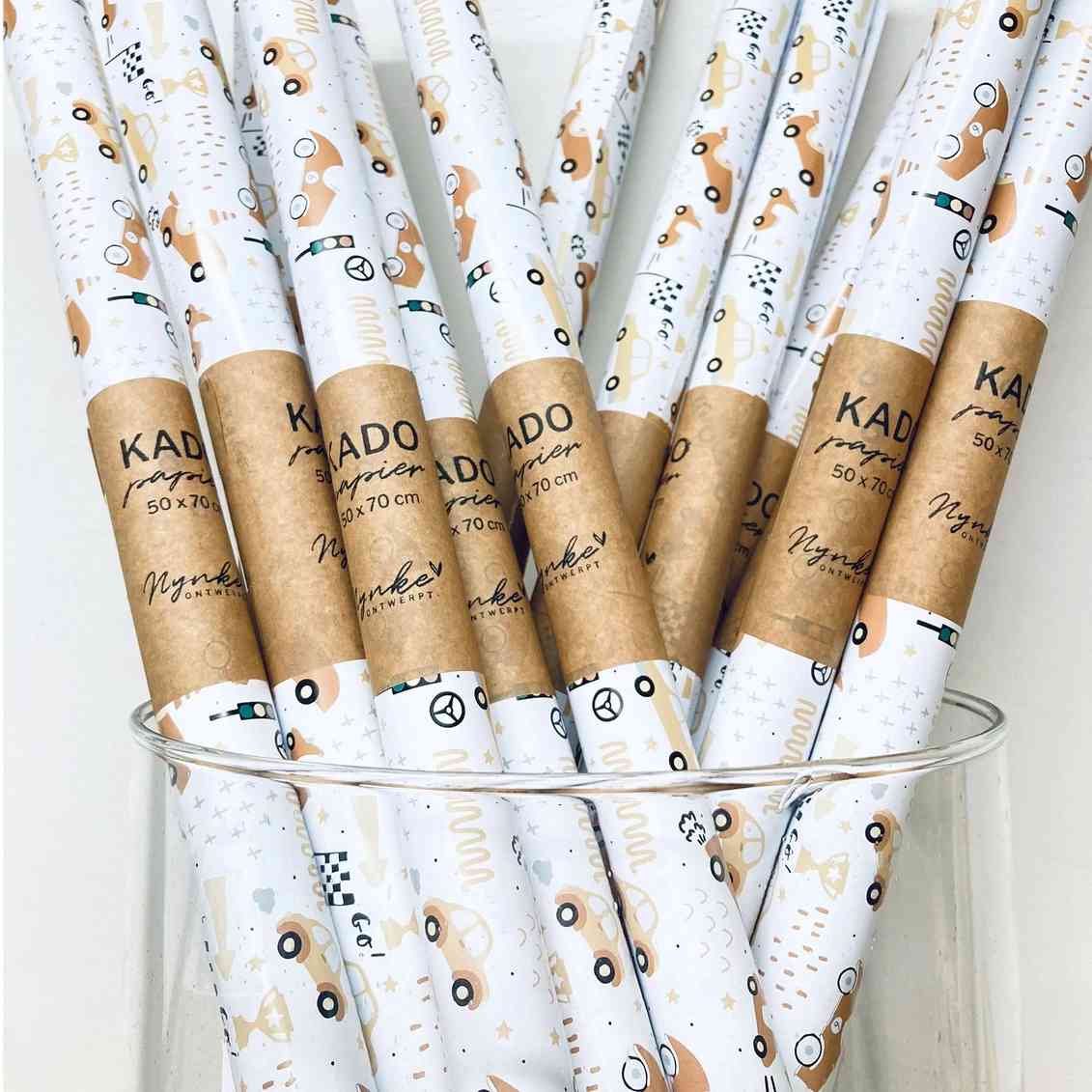 Eine Sammlung gerollter Geschenkpapiere wird in einem Glasbehälter präsentiert. Die Papiere haben einen weißen Hintergrund mit einem verspielten Muster aus kleinen Oldtimern und geschwungenen Linien in gedämpften Braun- und Grüntönen. Jede Rolle, perfekt für Geschenkpapier junge, ist mit einem Papierband mit der Aufschrift „Nynke Ontwerpt – Geschenkpapier Autos“ und Größenangaben von Nynke Ontwerpt beschriftet.