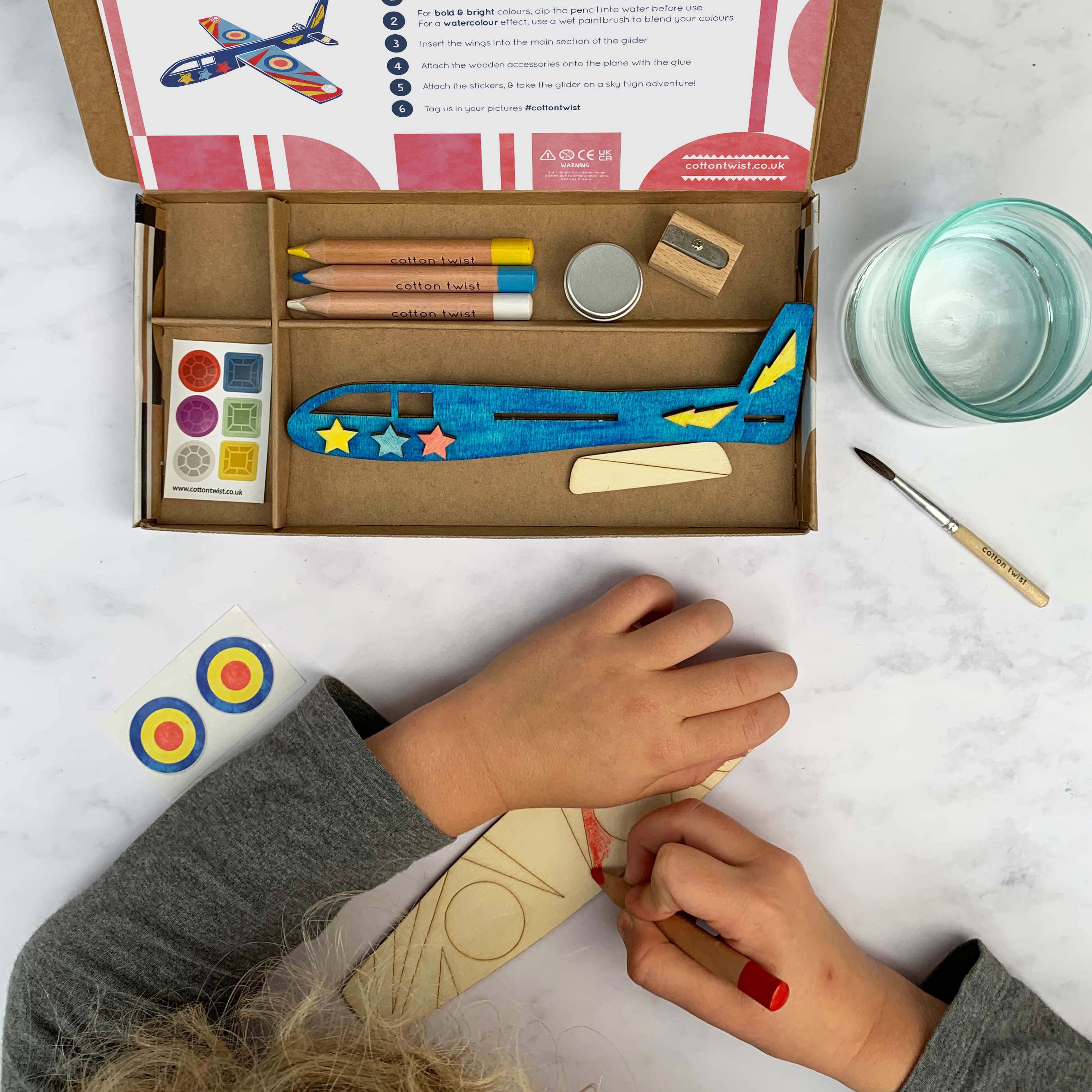 Eine Person dekoriert ein Holzflugzeug mit der Cotton Twist - DIY Flugzeug-Set Activity Box, die Buntstifte, Farbe, einen Pinsel, Aufkleber und eine Schritt-für-Schritt-Anleitung enthält. Die Cotton Twist Activity Box steht offen auf einem Tisch, verschiedene Materialien sind ordentlich in Fächern angeordnet.