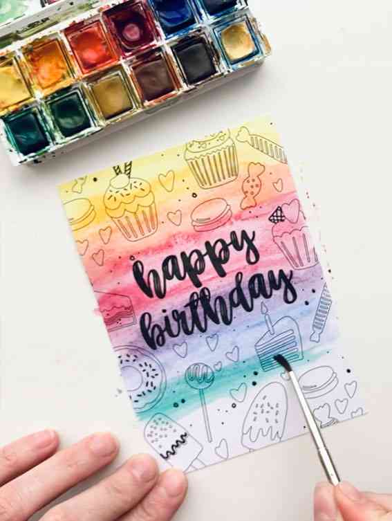 Eine farbenfrohe, handgemachte Nynke Ontwerpt - Karte Malkarte Süßes zeigt handgezeichnete Cupcakes, Herzen und Süßigkeiten mit „Happy Birthday“ in Schreibschrift auf einem Regenbogen-Aquarell-Hintergrund. Die Karte wird bemalt, wobei ein Pinsel und eine Palette mit Aquarellfarben im Bild sichtbar sind.