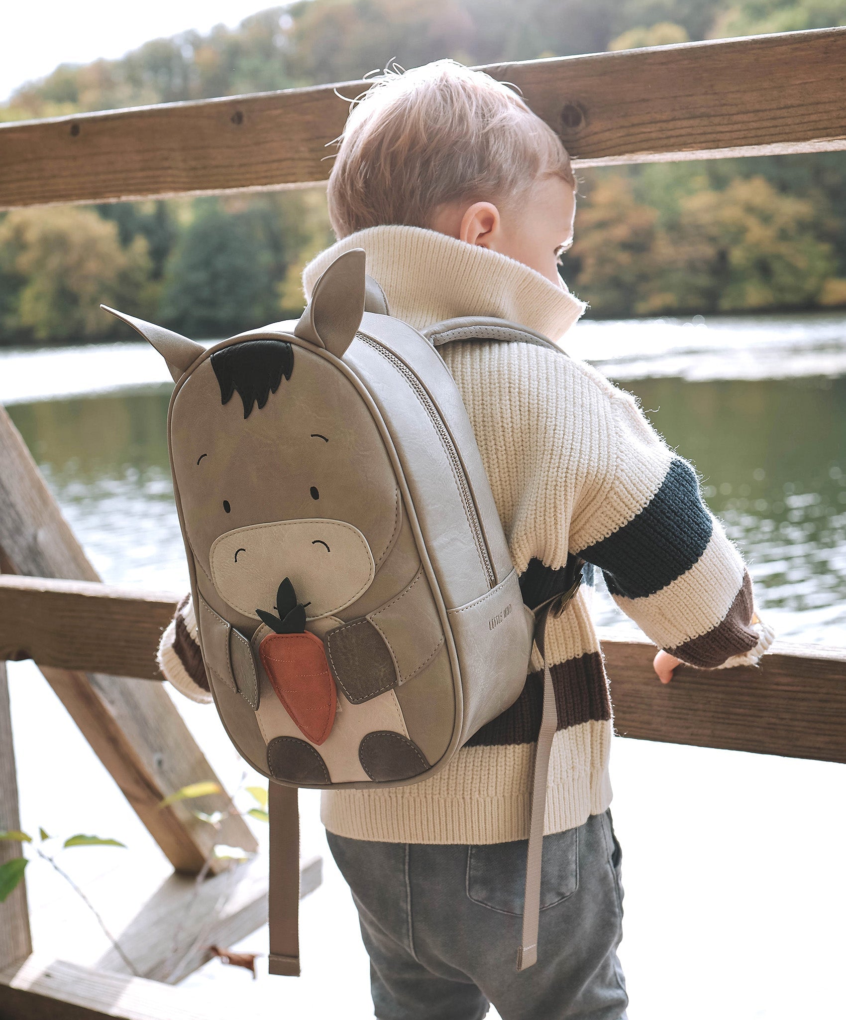 Ein kleines Kind steht auf einer Holzbrücke mit Blick auf einen von Bäumen umgebenen See. Es trägt einen kuscheligen Pullover und einen skurrilen Rucksack von Little Who – Rucksack Esel Edvin in Form eines Pferdes mit Karottenmuster. Das von hinten aufgenommene Kind scheint in Gedanken versunken, während seine nachhaltig hergestellte Kleidung die ruhige Naturszene ergänzt.