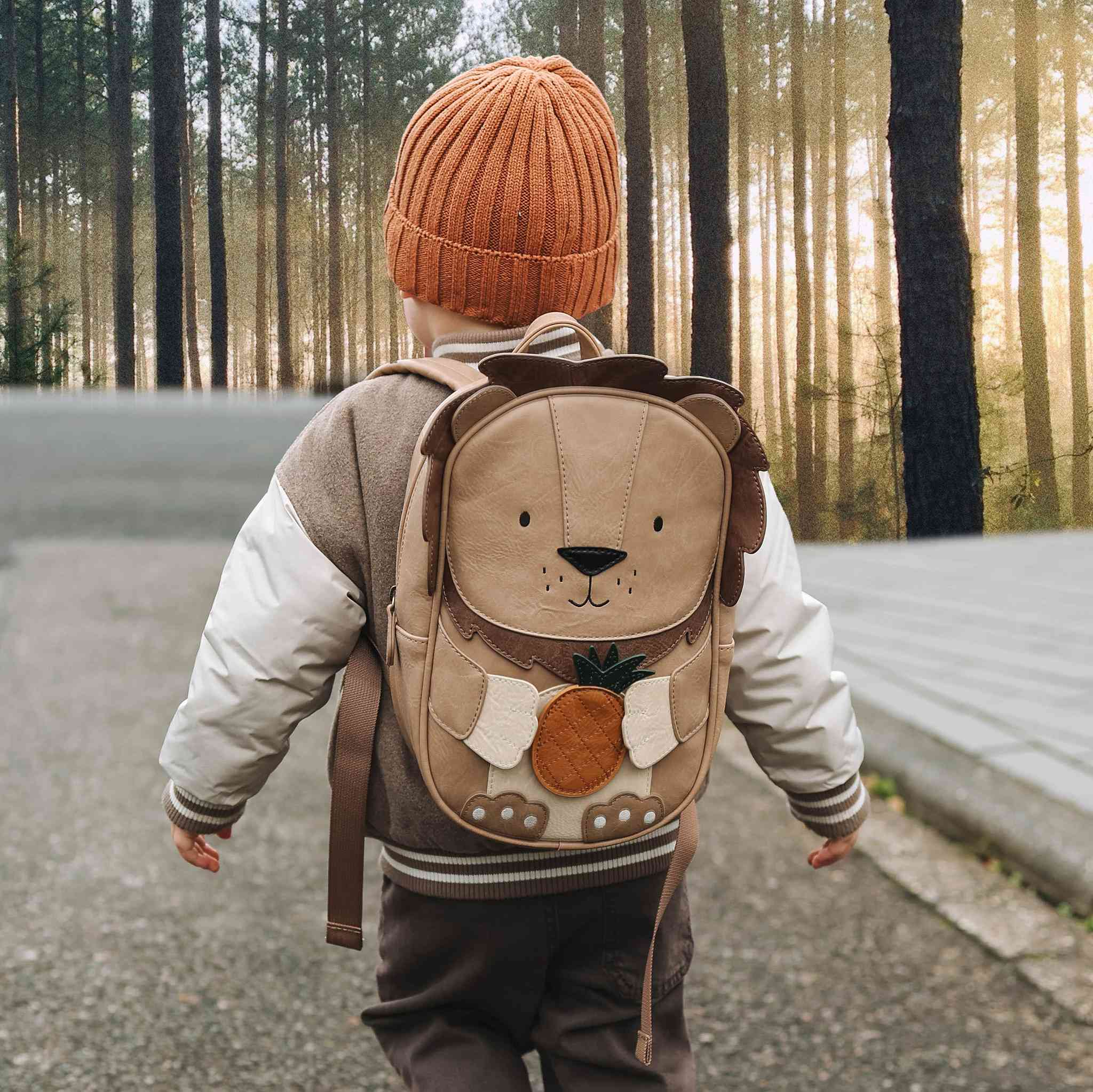 Ein Kind mit einer rotbraunen Strickmütze und einer beigen Jacke mit weißen Ärmeln läuft auf einem gepflasterten Weg durch einen Wald. Das Kind hat einen süßen Little Who - Rucksack Löwe Kaius dabei, der mit spülmaschinenfesten Aufklebern verziert ist, auf denen kleine orangefarbene Karottenmotive abgebildet sind. Sonnenlicht fällt durch die hohen Bäume im Hintergrund.