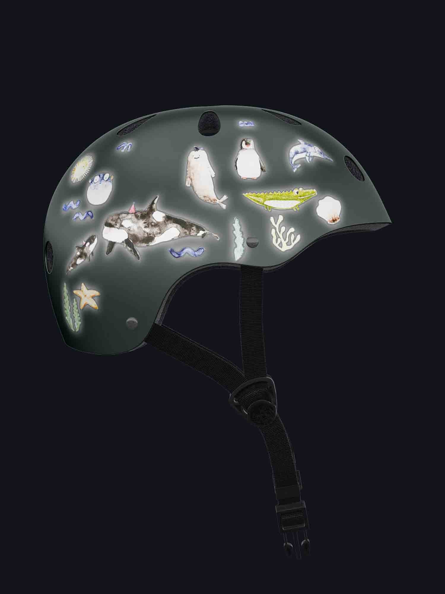 Ein dunkelfarbiger Helm zeigt verschiedene leuchtende Illustrationen, darunter einen Hai, Pinguine, einen Seestern, ein Fossil und Wellen vor schwarzem Hintergrund. Die Helmriemen hängen darunter und das Gesamtdesign ist skurril und ozeanisch – perfekt für jedes Baby oder Kleinkind, das seine ersten Abenteuer im Freien mit Halfbird genießt – Set Reflektierende Aufkleber: Meereswelt von Halfbird.
