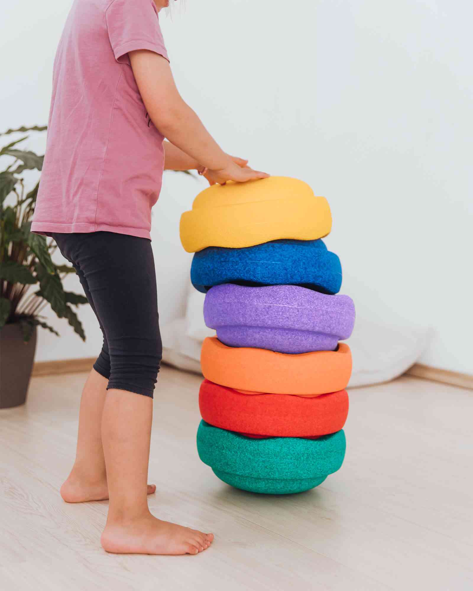 Eine Person in einem rosa Shirt und schwarzen Leggings stapelt bunte Kissen oder Trittsteine von Stapelstein - Rainbow classic (6 Stück) von Stapelstein in absteigender Reihenfolge von gelb, blau, lila, rot, orange und grün. Im Hintergrund ist eine grüne Pflanze auf einem hellen Holzboden zu sehen.