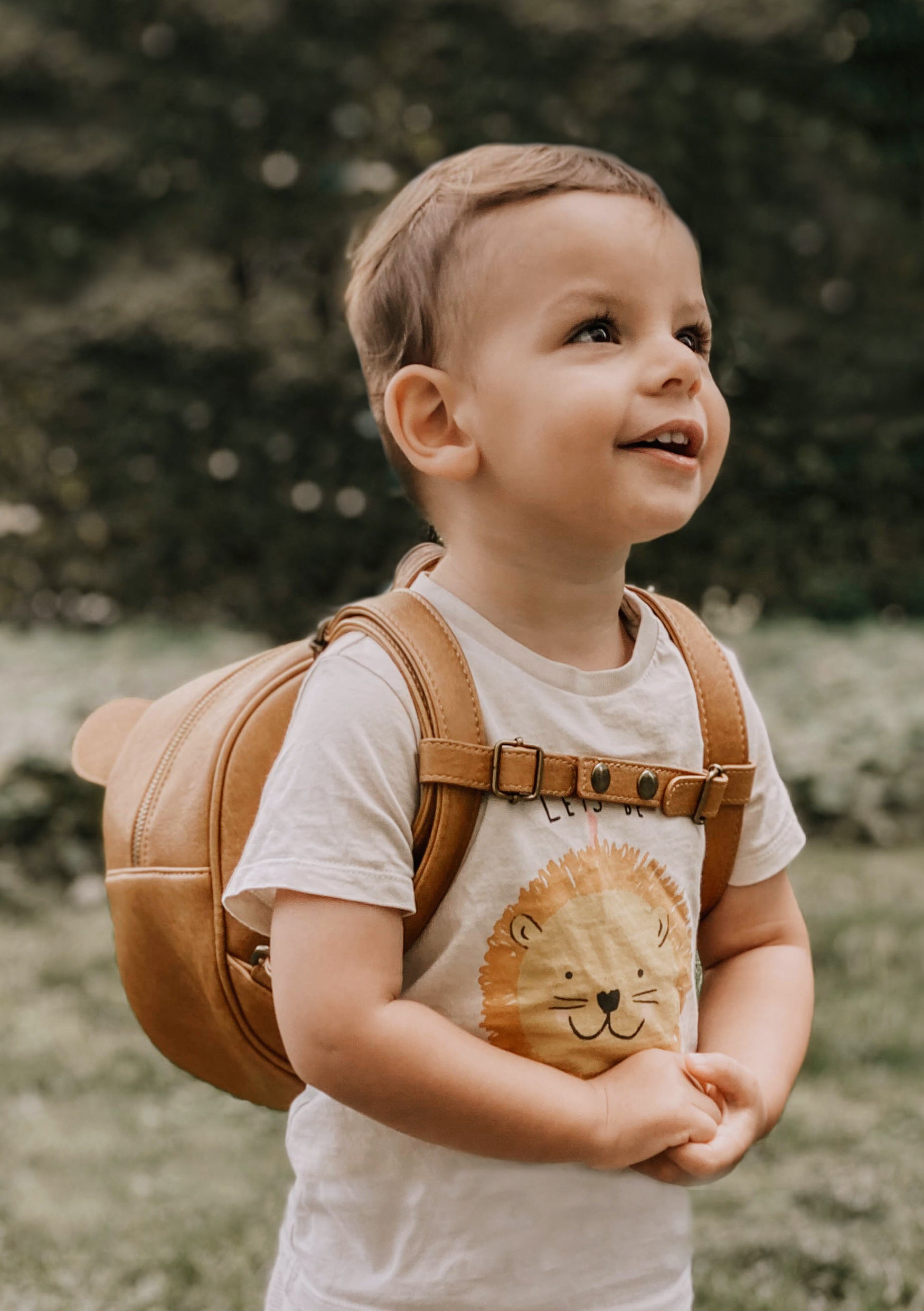 Ein kleines Kind mit kurzen hellbraunen Haaren lächelt im Freien. Das Kind trägt ein weißes T-Shirt mit einem Löwenmotiv und einen braunen Rucksack mit einem Little Who-Aufkleber – Brustgurt von Little Who. Der Hintergrund ist verschwommenes Grün.